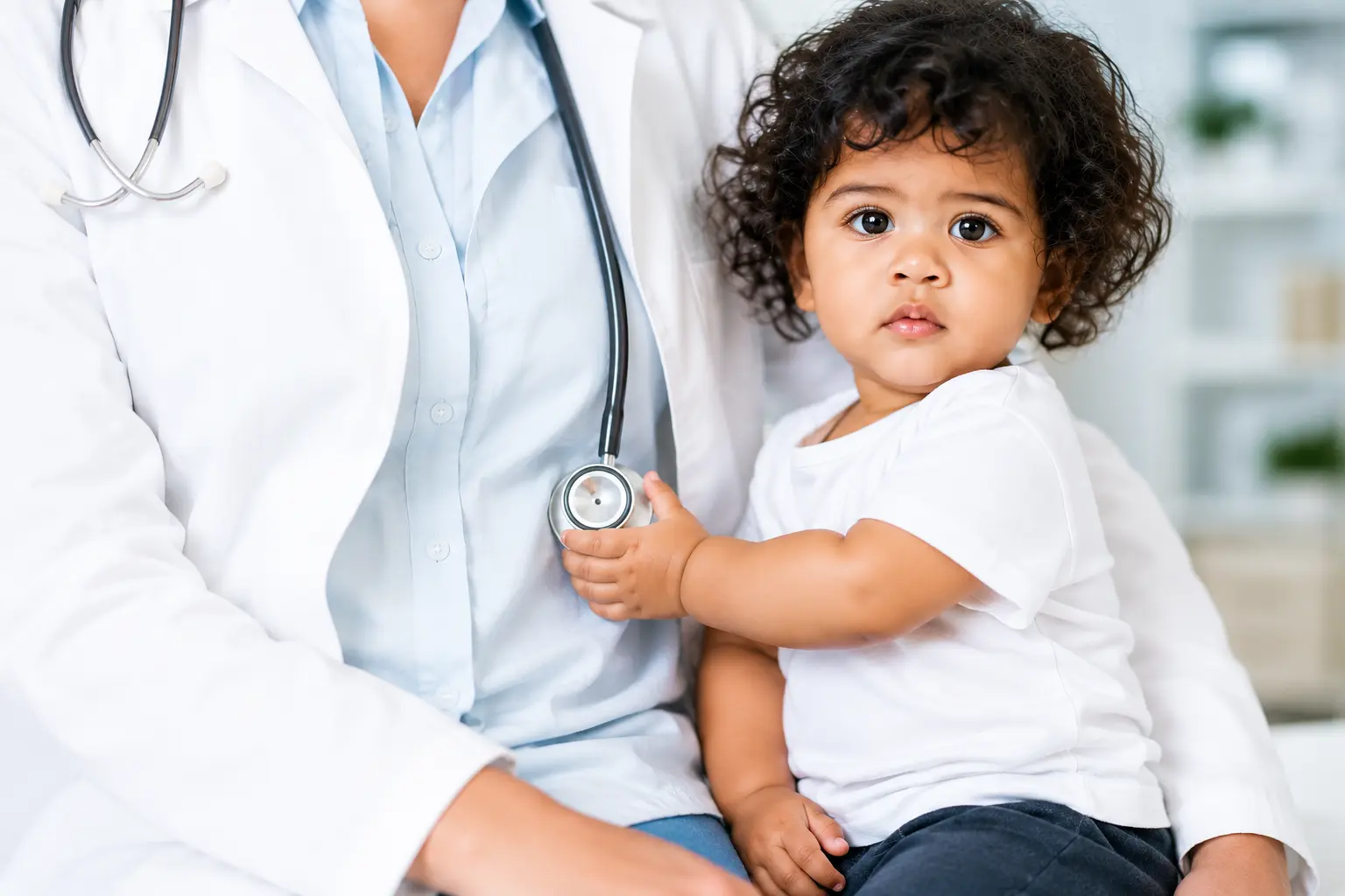 “Cute toddler sitting on a doctor’s lap holding a stethoscope in a bright, clean pediatric clinic, representing child-friendly healthcare and medical care for children.”